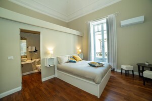 Una camera da letto con un letto, comodini, una finestra con tende, un tavolino e un bagno visibile attraverso una porta aperta.
