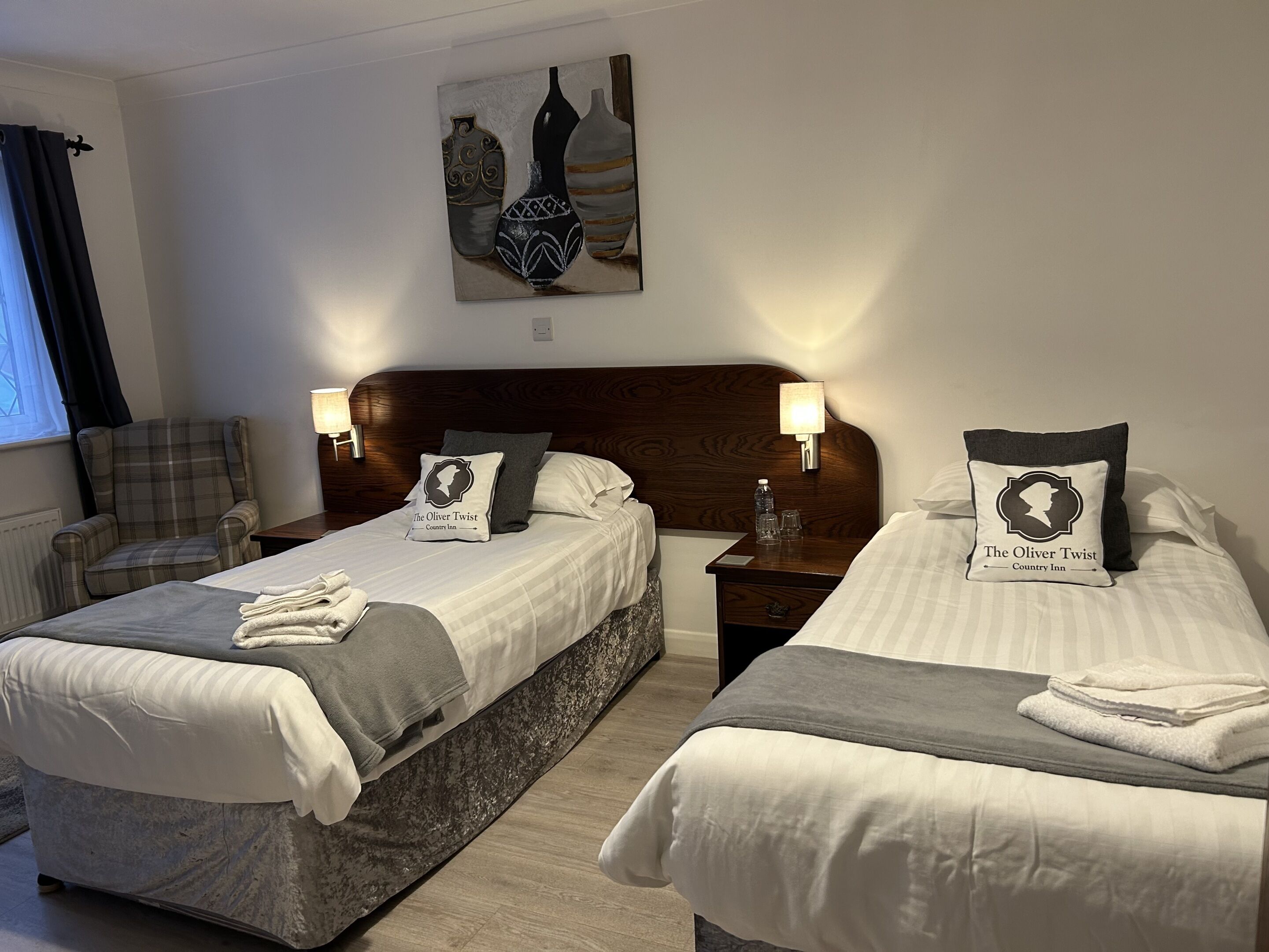 A hotel room with two beds, a headboard, bedside lamps, a painting, and a window with curtains.