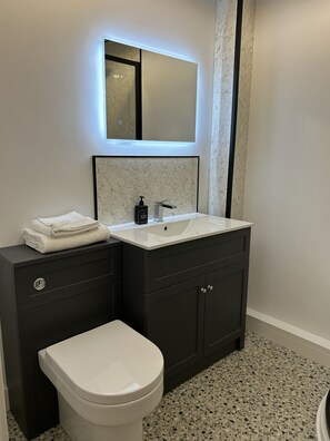 Un bagno moderno con un water bianco, mobili antracite, un lavandino in ceramica con schienale in marmo e uno specchio a parete illuminato da luci a LED sotto il mobile.