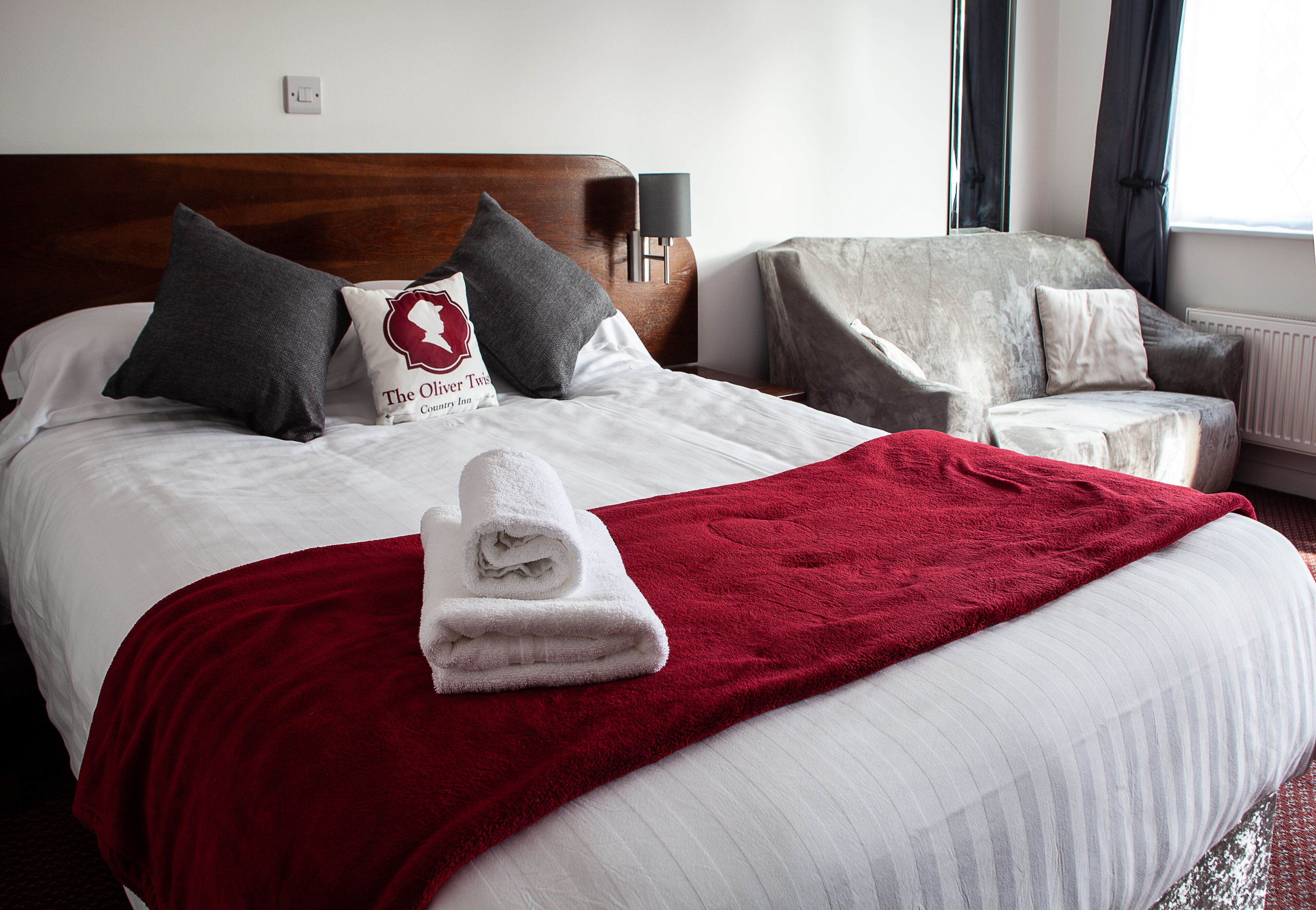 Una cama bien hecha con una colcha roja, una toalla blanca doblada, dos almohadas grises y una almohada con un logotipo.