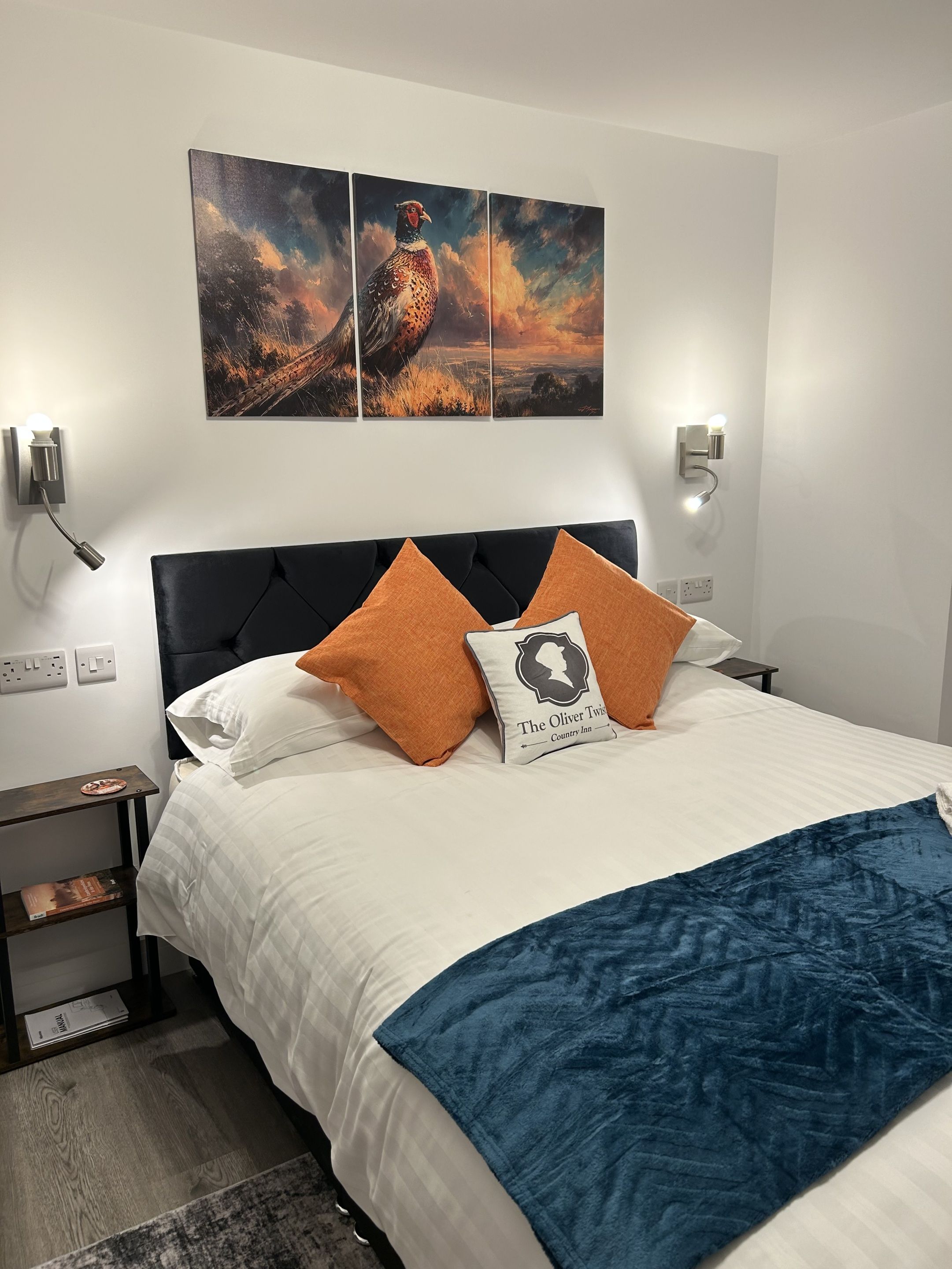 A neatly made bed with a white and blue blanket, two orange pillows, and a pillow with a silhouette design. Above the bed, there is a three-panel artwork depicting a bird.
