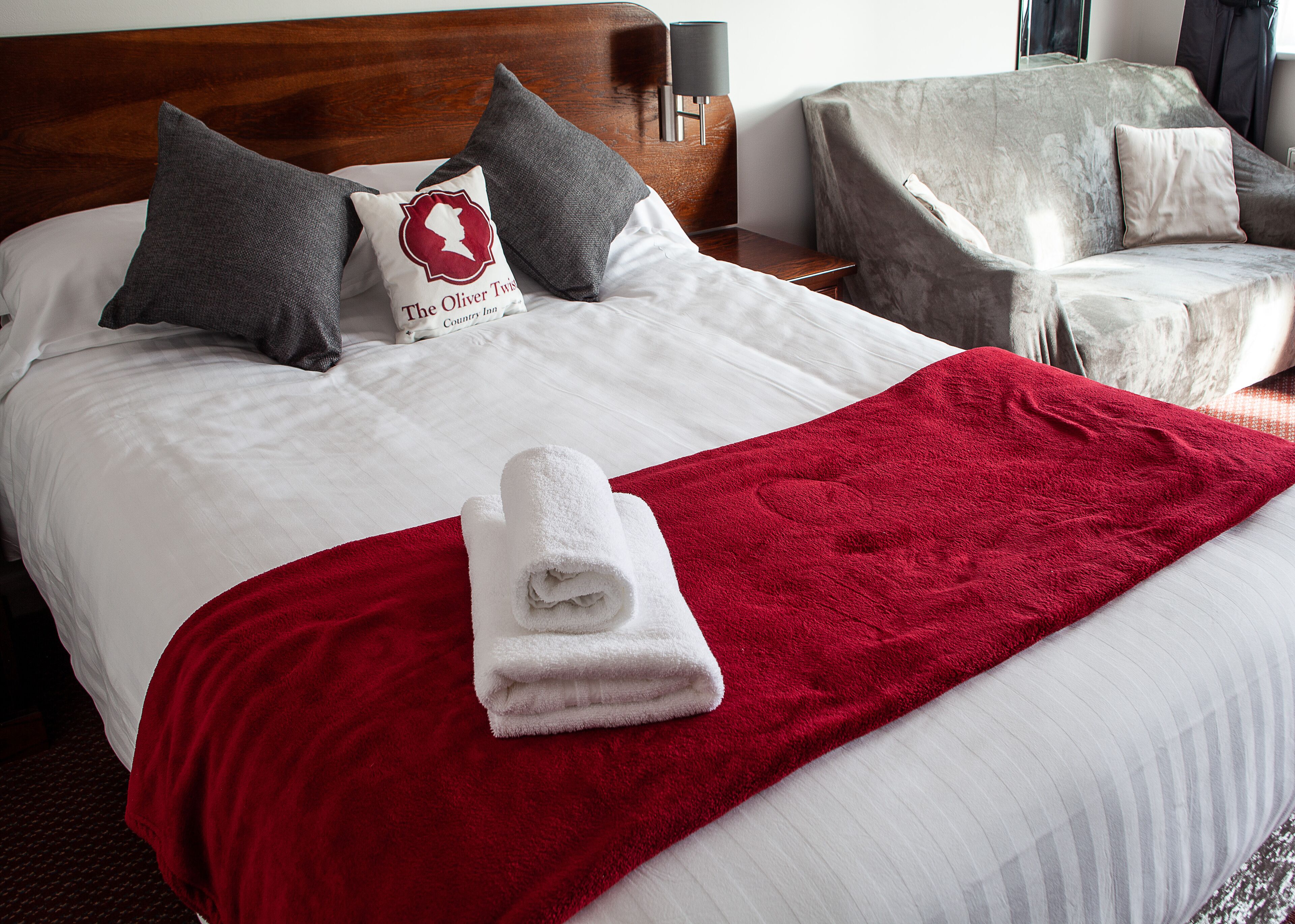 A neatly made bed with a red blanket and a folded white towel, two pillows, a nightstand with a lamp, and a grey armchair.