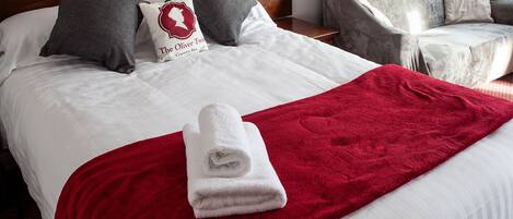 A neatly made bed with a red blanket and a folded white towel, two pillows, a nightstand with a lamp, and a grey armchair.