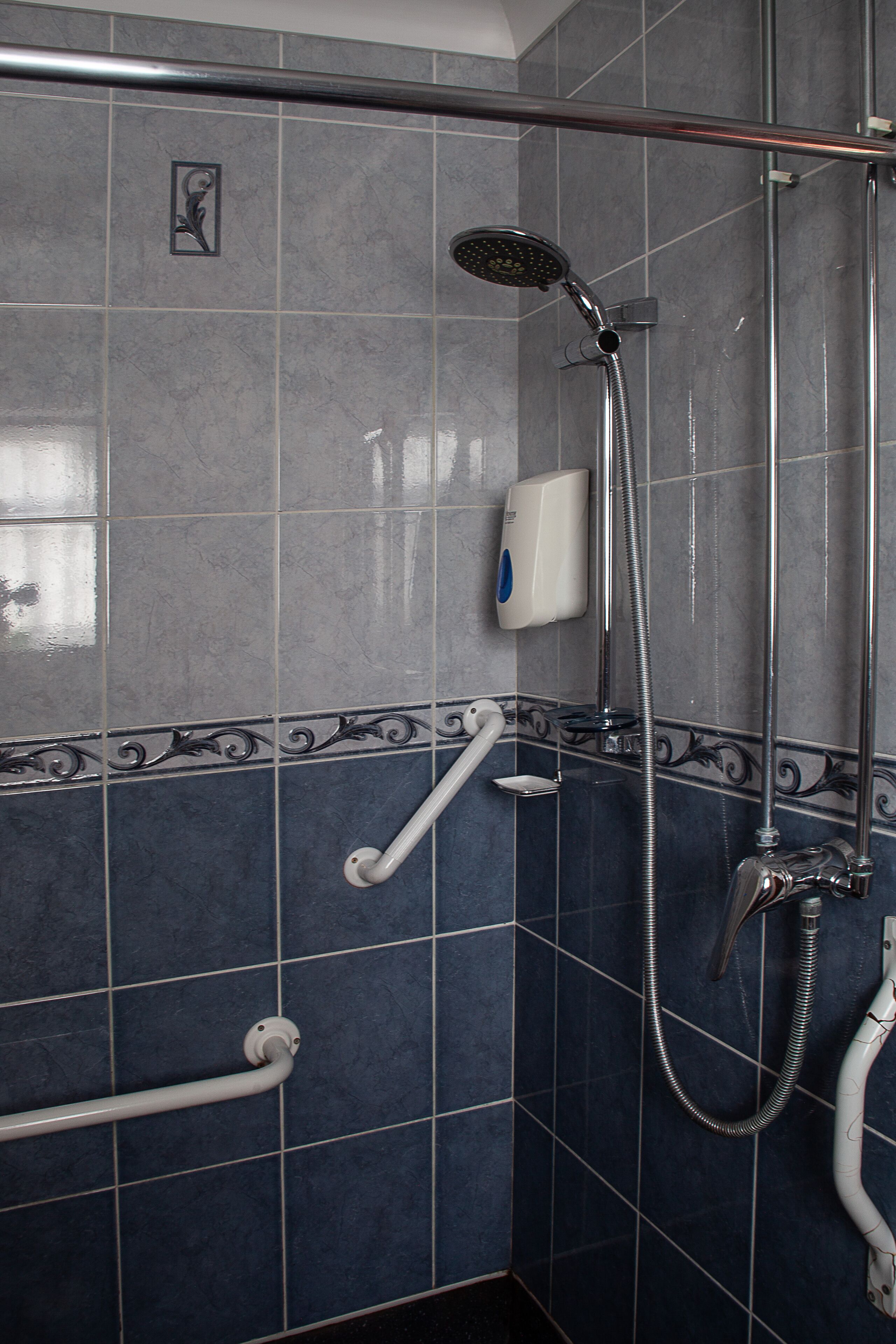 A bathroom with blue tiles, a shower with a handheld showerhead, a handheld showerhead, a soap dispenser, and a grab bar.
