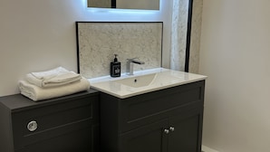 A modern bathroom with a white toilet, dark grey cabinets, a white sink with a marble backsplash, and a wall-mounted mirror with under-cabinet lighting. - Oliver Twist Country Inn (Wisbech)