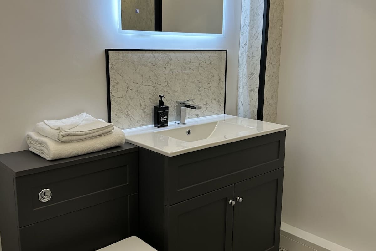 A modern bathroom with a white toilet, dark grey cabinets, a white sink with a marble backsplash, and a wall-mounted mirror with under-cabinet lighting.