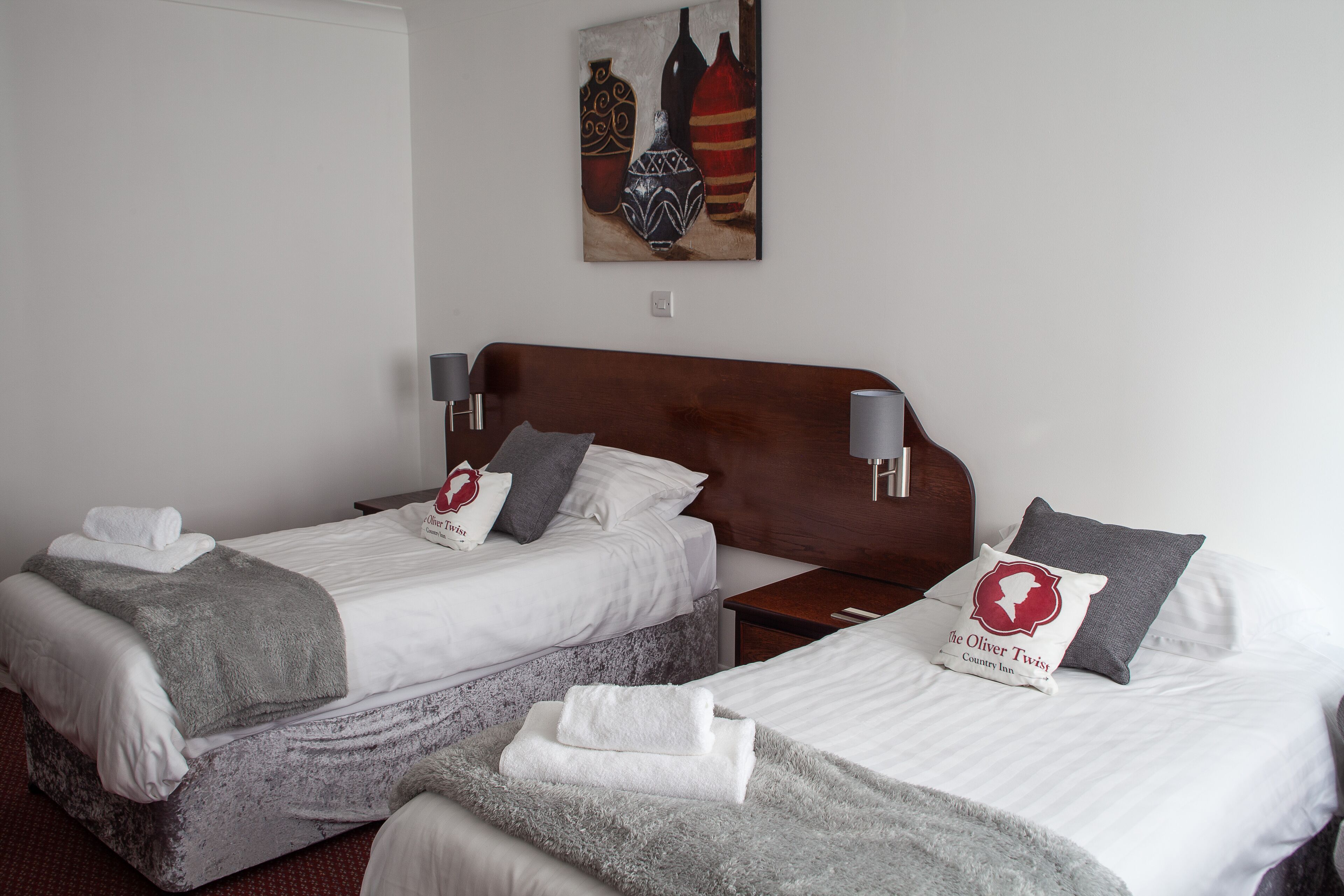 A hotel room with two beds, each with white linens and gray blankets. There are decorative pillows and a painting of vases on the wall.