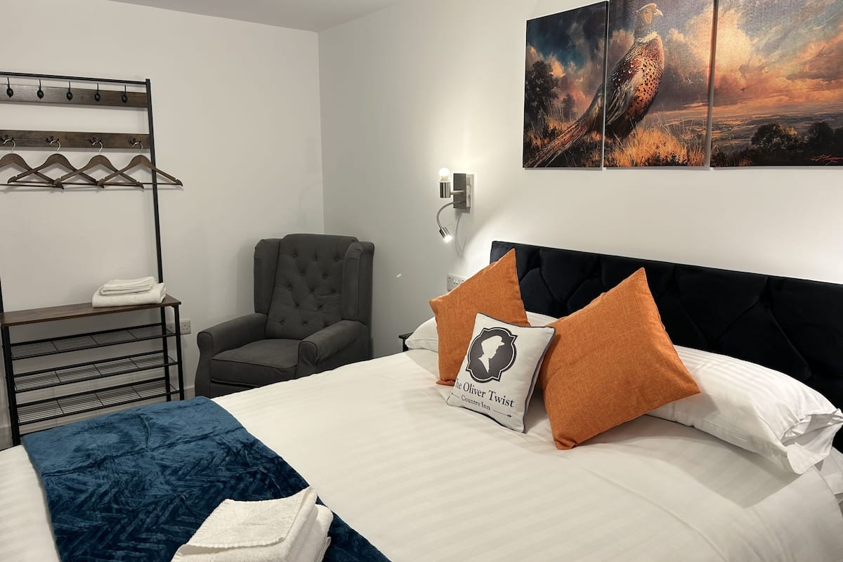 A hotel room with a bed, a chair, a coat rack, and a wall-mounted light.