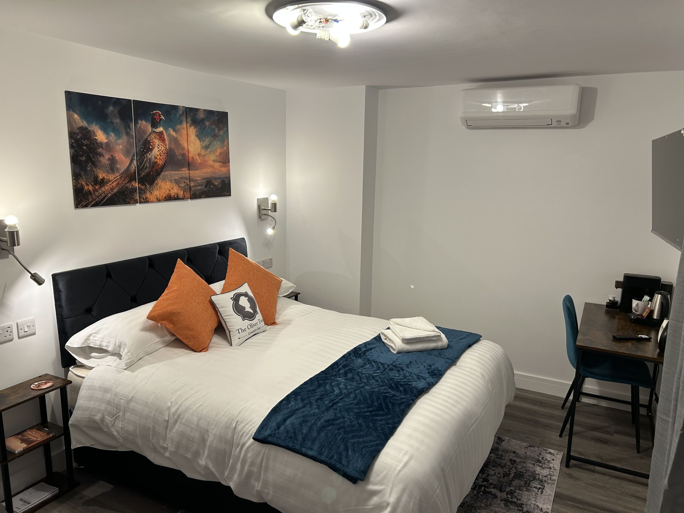 Ett hotellrum med en säng, ett skrivbord och en stol. Det finns en tavla på väggen och en luftkonditioneringsanläggning ovanför skrivbordet.