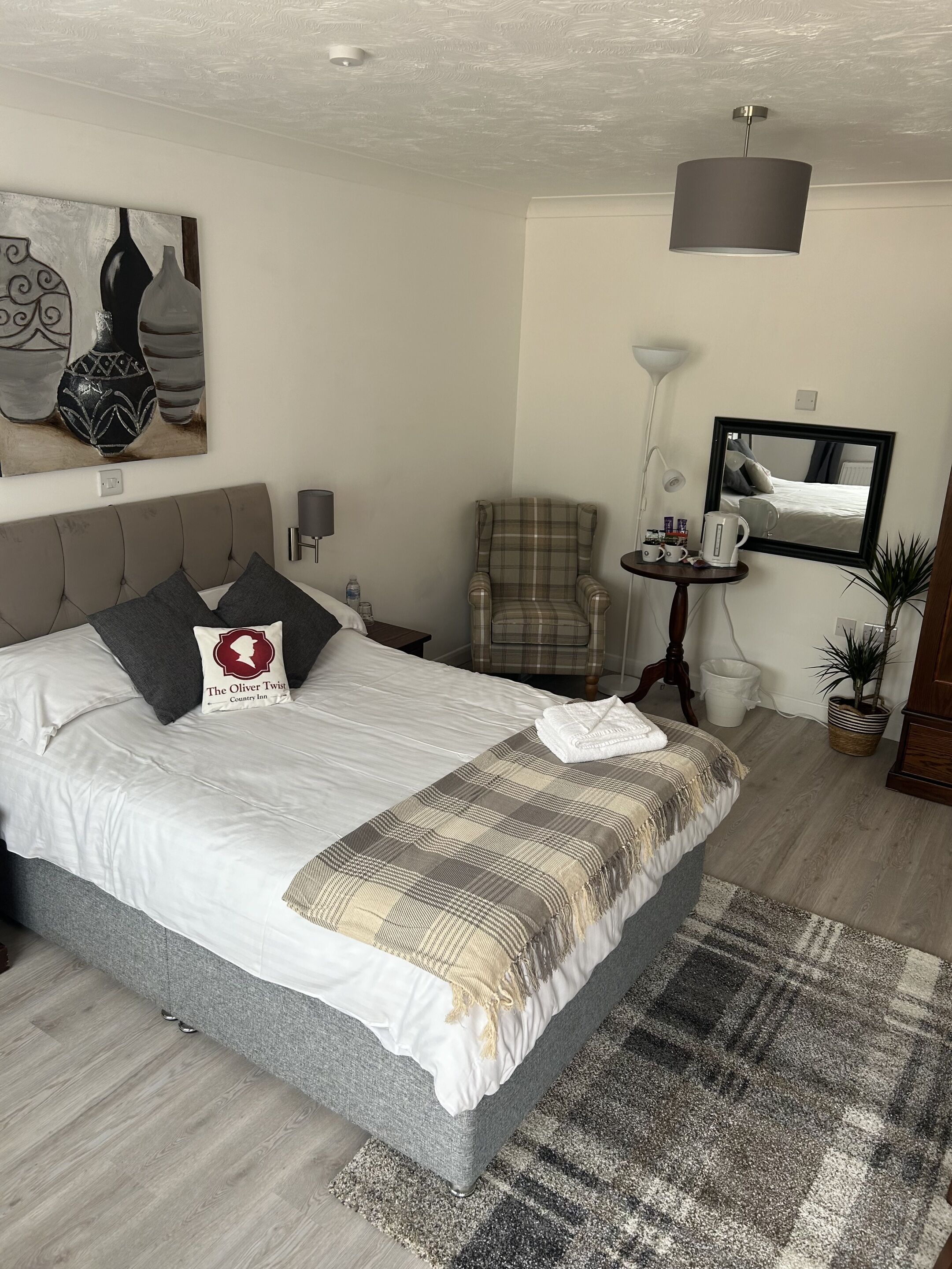 A neatly arranged bedroom with a large bed, a chair, a small table with a kettle, a mirror, and a painting on the wall.