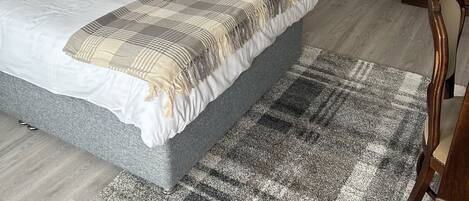 A neatly arranged bedroom with a large bed, a plaid armchair, a small table with a lamp, and a mirror on the wall.