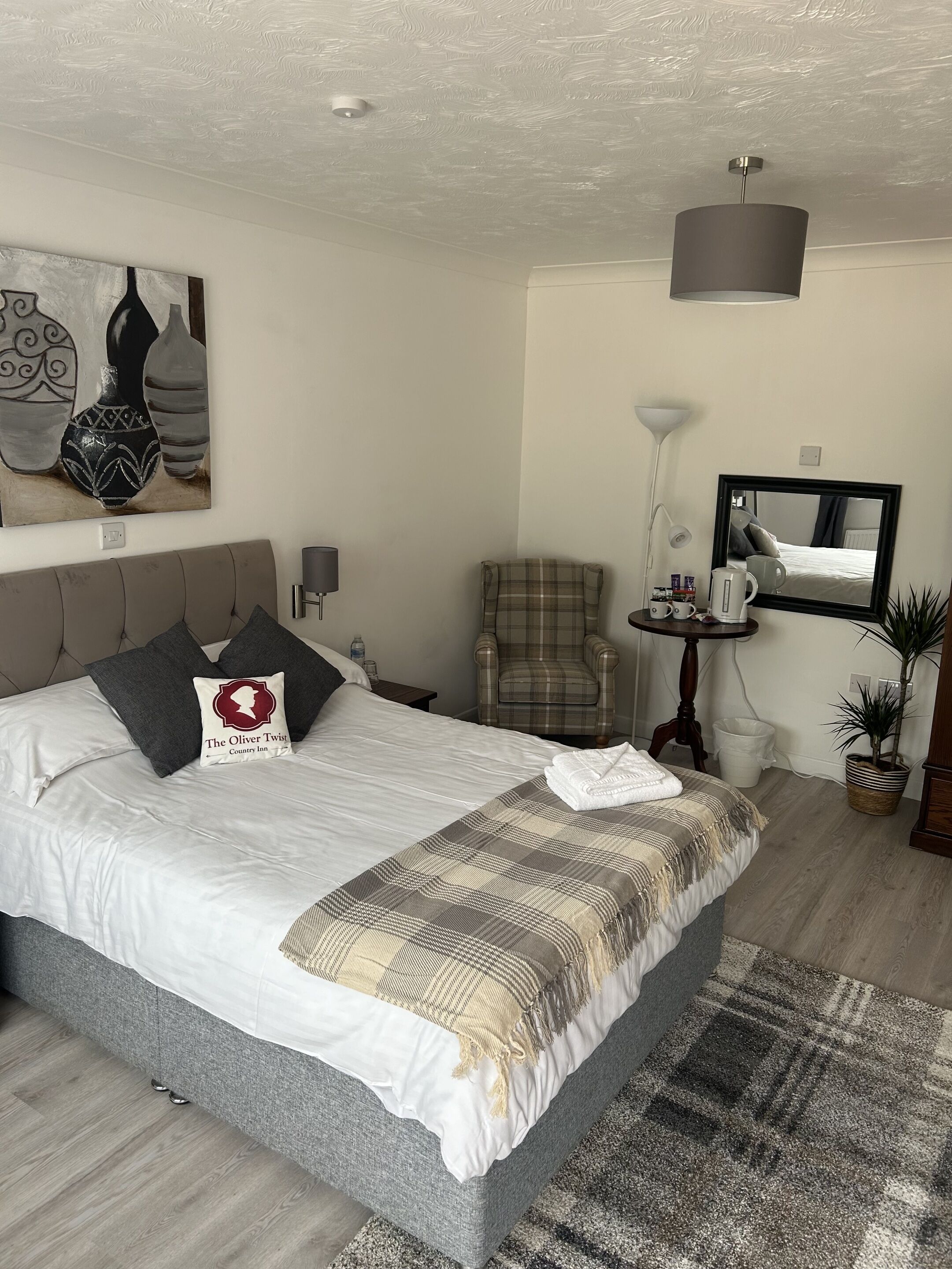 A neatly made bed with a plaid blanket, a plaid chair, a small table with a lamp, and a painting on the wall.