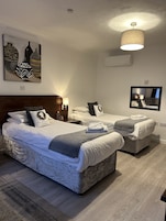Habitación de hotel con una cama grande, dos camas individuales, cabecera, mesitas de noche, lámpara, espejo y un cuadro en la pared.