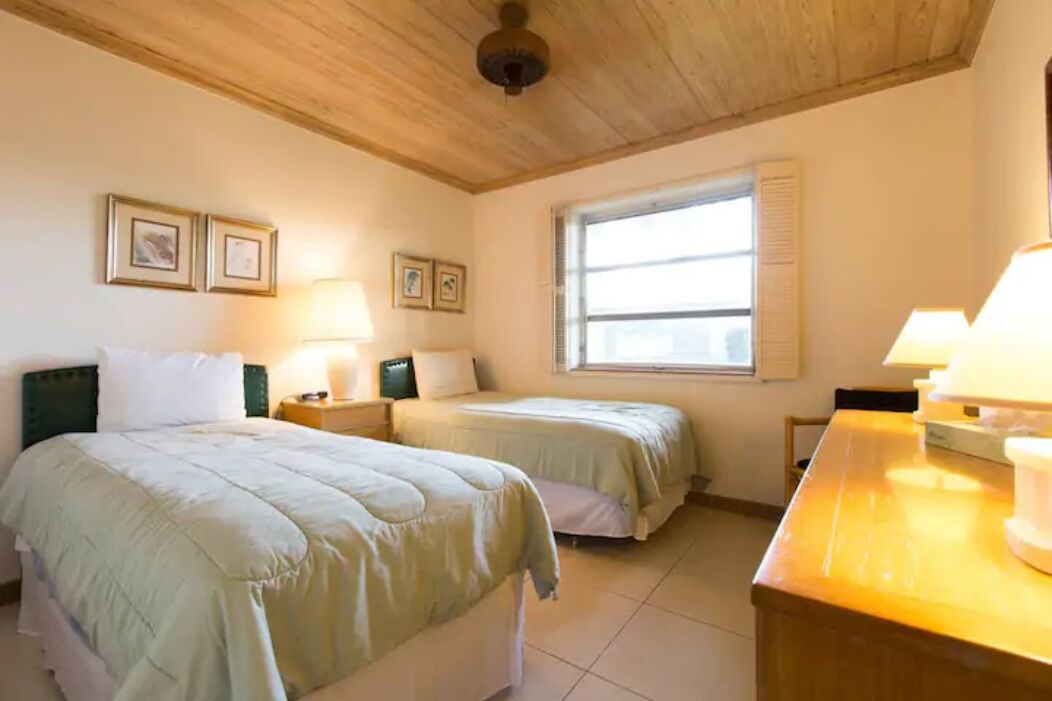 A room with two beds, a wooden ceiling, framed pictures on the wall, a window with blinds, and a table with a lamp.