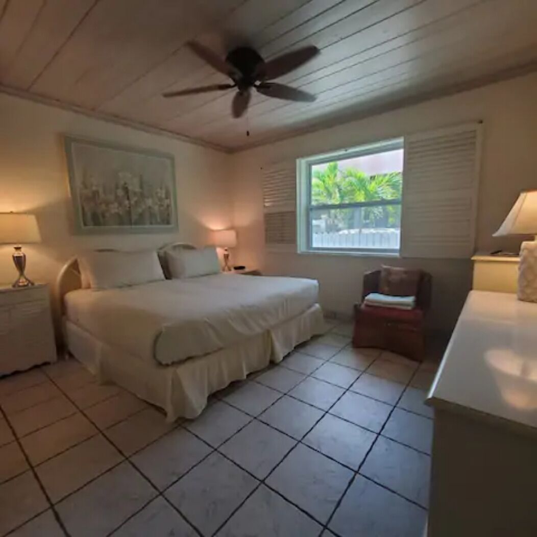 A bedroom with a bed, a ceiling fan, a window with a view, and a small table with a lamp.