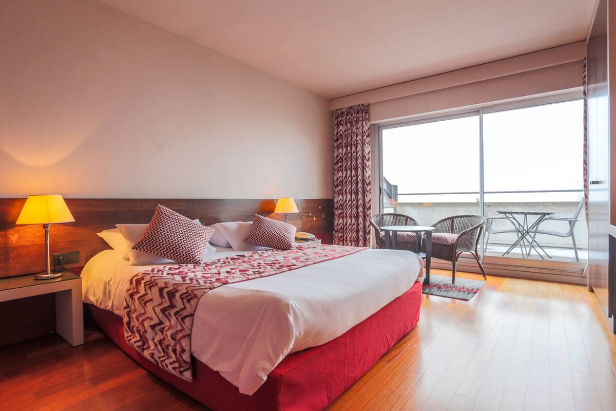 A hotel room with a large bed, two bedside lamps, a wooden headboard, a balcony with a table and chairs, and a view of the sky.