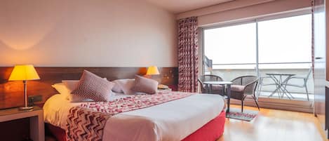 A hotel room with a large bed, two bedside lamps, a wooden headboard, a balcony with a table and chairs, and a view of the sky.