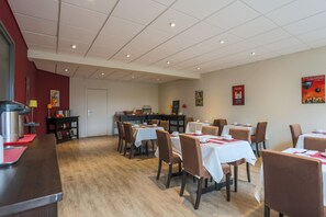 A dining area with tables and chairs, a kitchen in the background, and a red wall with artwork.