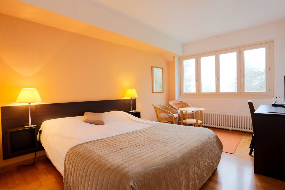 A hotel room with a large bed, two bedside lamps, a desk, a chair, and a window with blinds.