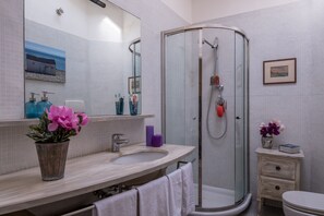 Une salle de bain moderne avec une douche à parois de verre, un lavabo avec un miroir et une petite table de chevet agrémentée d’un vase de fleurs.
