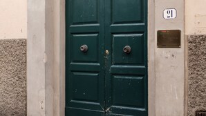 A green double door with a metal grille above it, set in a beige building with a stone base. A plaque with the number 21 is visible on the wall to the right of the door. - Santo Spirito Apartment (Florence)