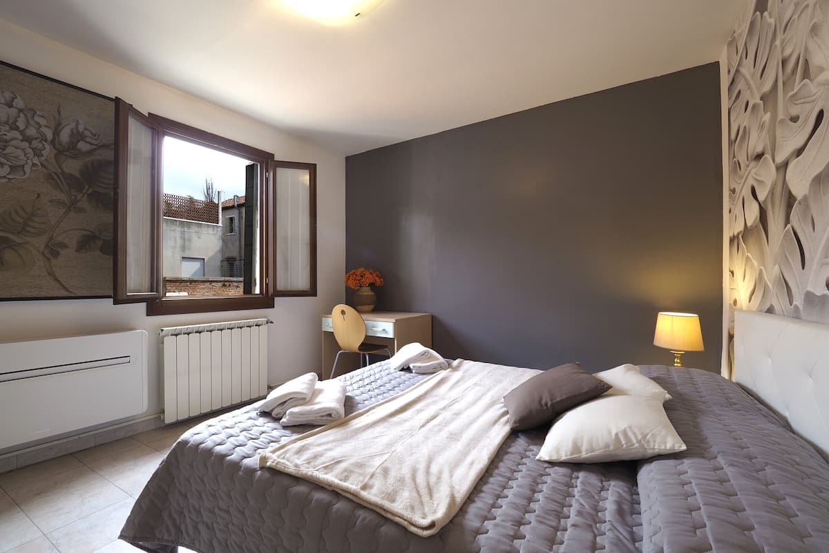 A bedroom with a bed, a desk, a chair, a radiator, a window, and a wall with a floral pattern.