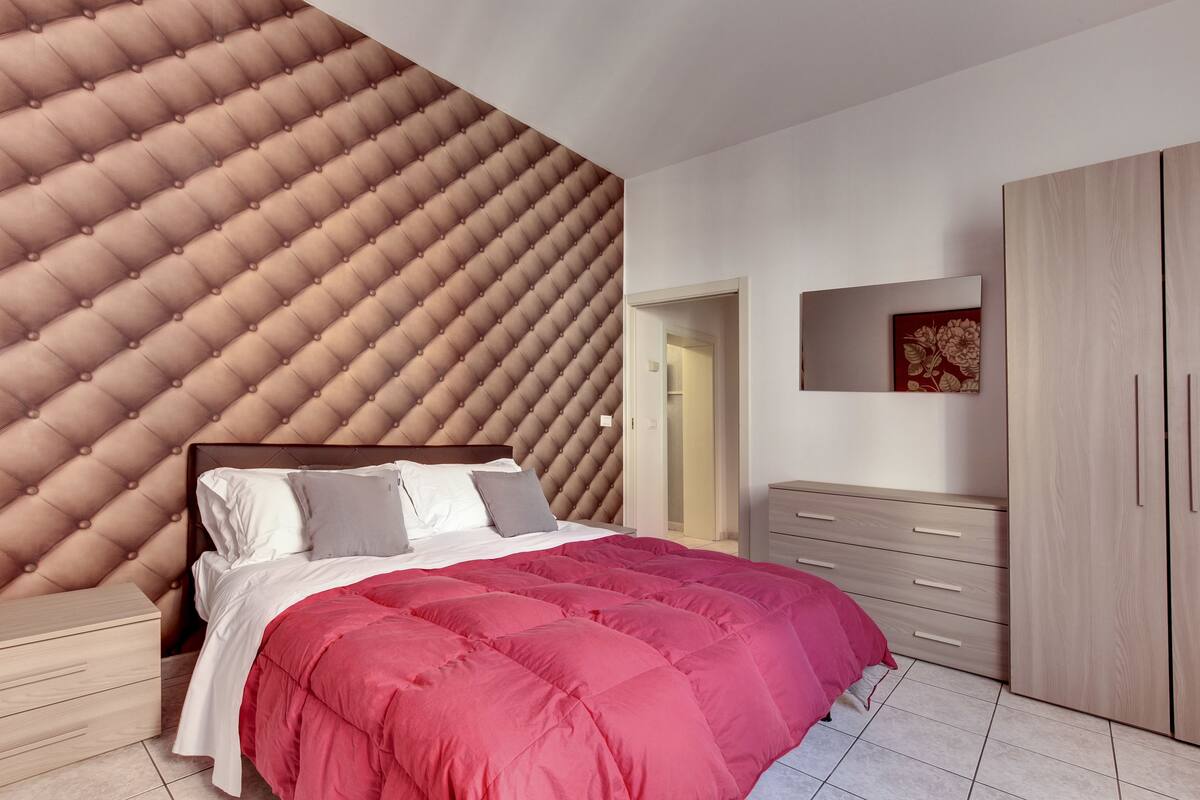 A bedroom with a tufted headboard, a bed with a red comforter, wooden nightstands, a wardrobe, and a mirror.