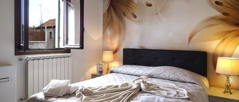 A bedroom with a bed, two bedside lamps, a radiator, and a window with an open shutter.