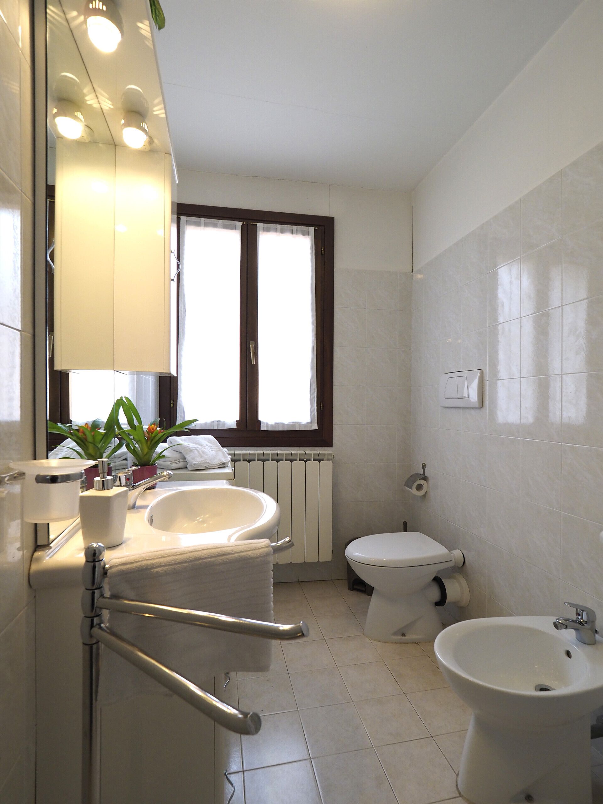 A modern bathroom with a large mirror, a sink, a toilet, and a bidet. There is a window with curtains, a radiator, and a towel rack.