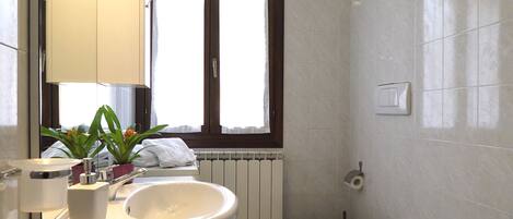 A modern bathroom with a large mirror, a sink, a toilet, and a bidet. There is a window with curtains, a radiator, and a towel rack.