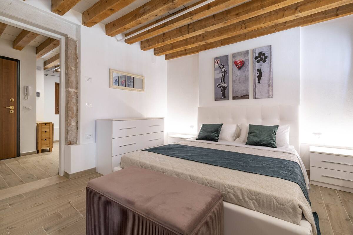 A bedroom with a bed, a bench, a dresser, and artwork on the wall.