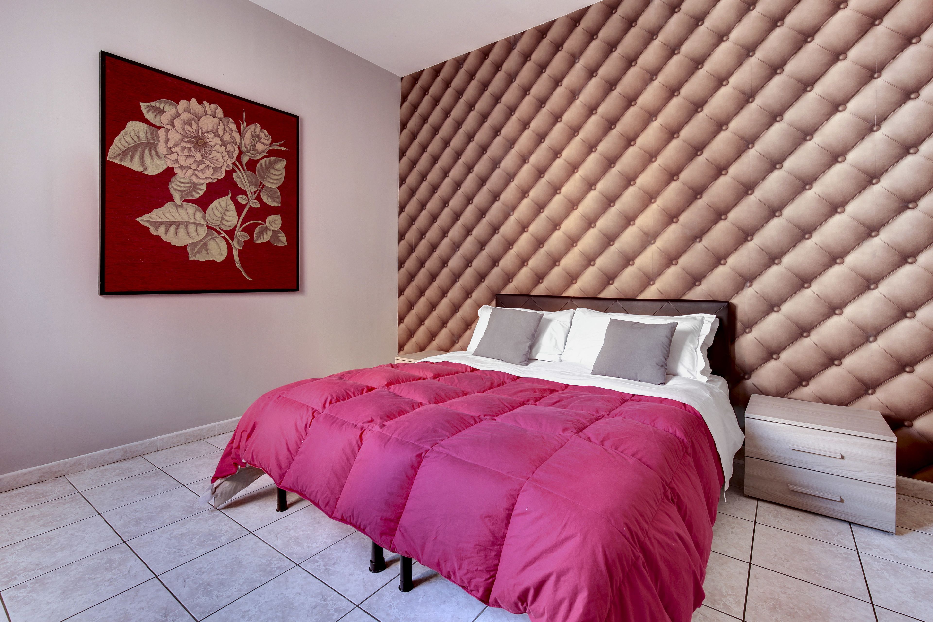 Ein Schlafzimmer mit einem gesteppten Kopfteil, einem Bett mit rosa Bettdecke, einem Nachttisch mit Schubladen und einem Blumenbild an der Wand.