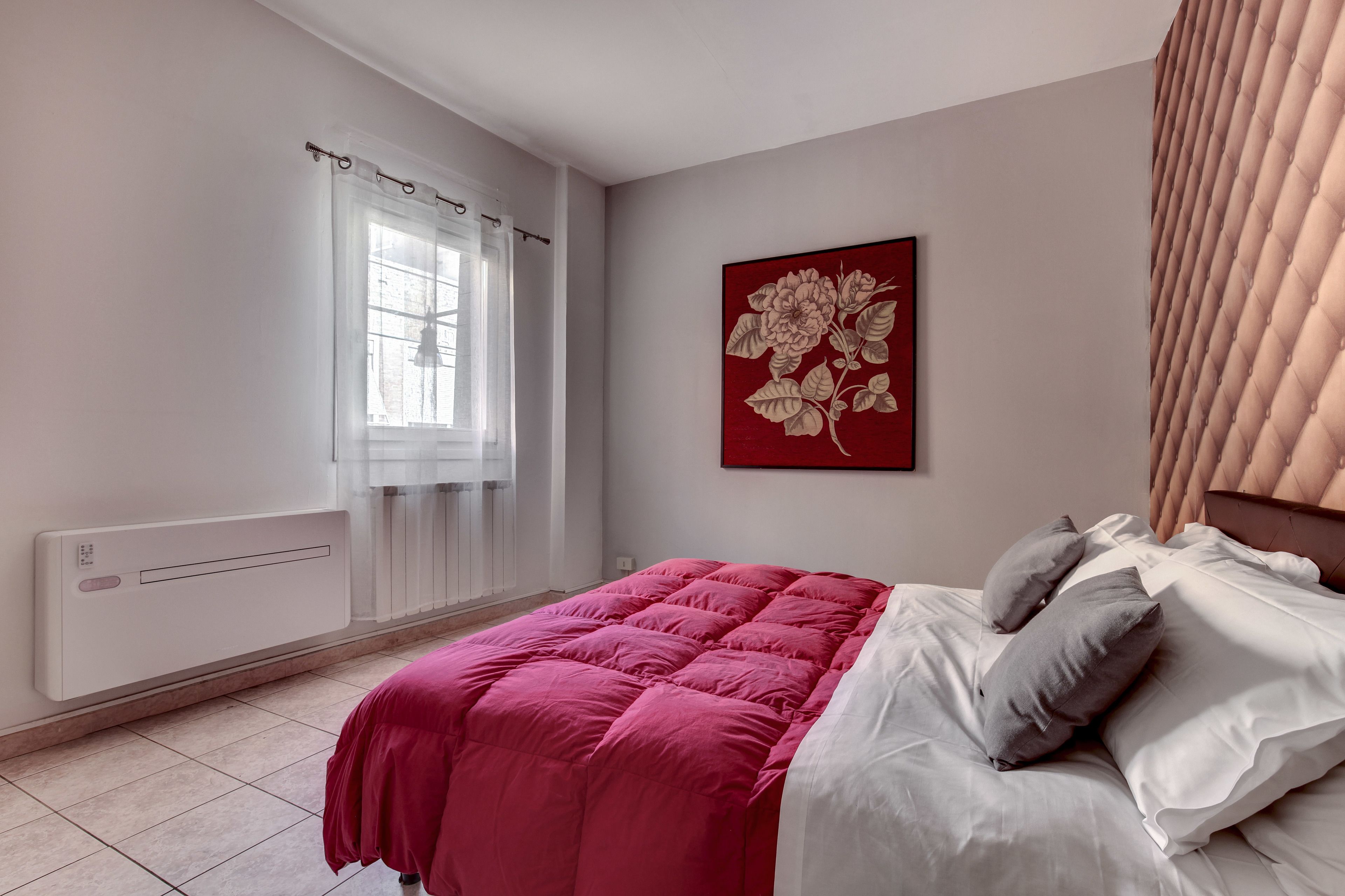 Un dormitorio con cama, radiador, ventana con cortinas y una obra de arte floral en la pared.