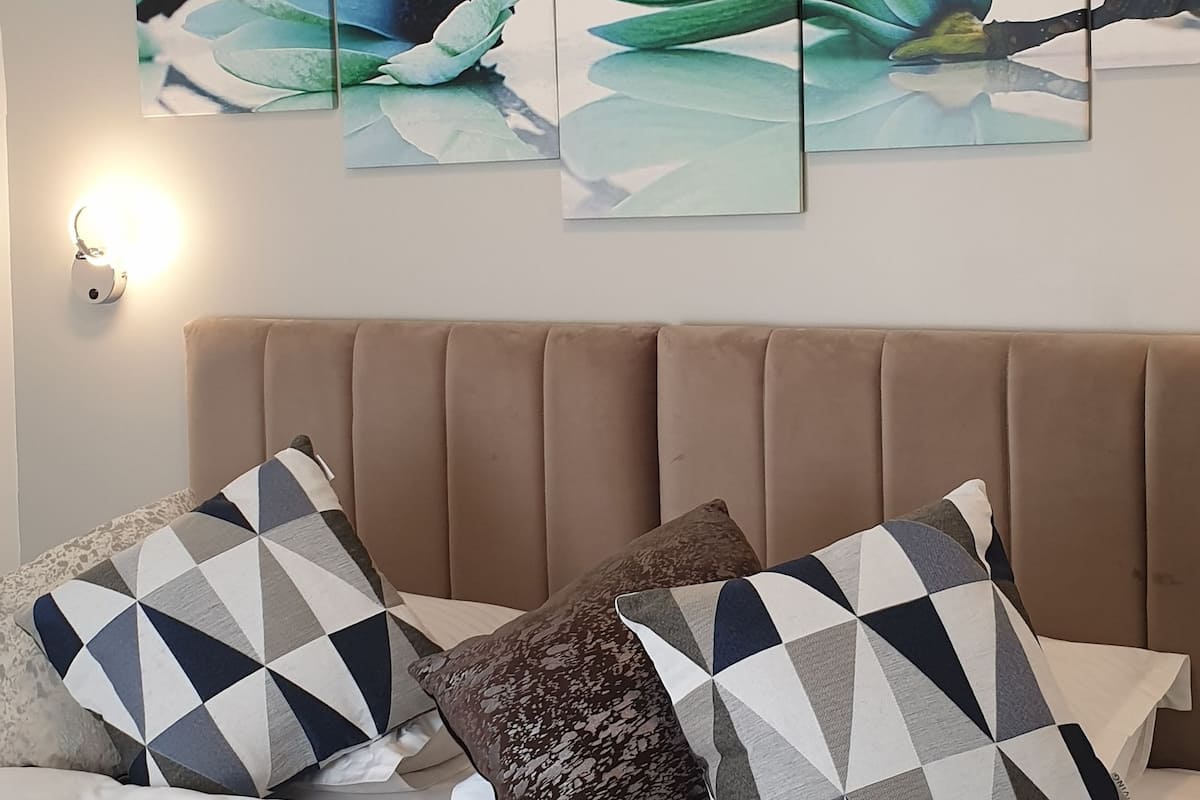 A neatly made bed with a brown headboard, a white blanket, and a folded white towel. There are three geometric patterned pillows and a brown textured pillow. Above the bed, there are three framed artworks depicting a flower.