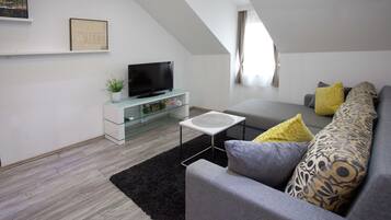A modern living room with a flat-screen TV on a glass cabinet, a grey sofa with patterned cushions, and a black rug.