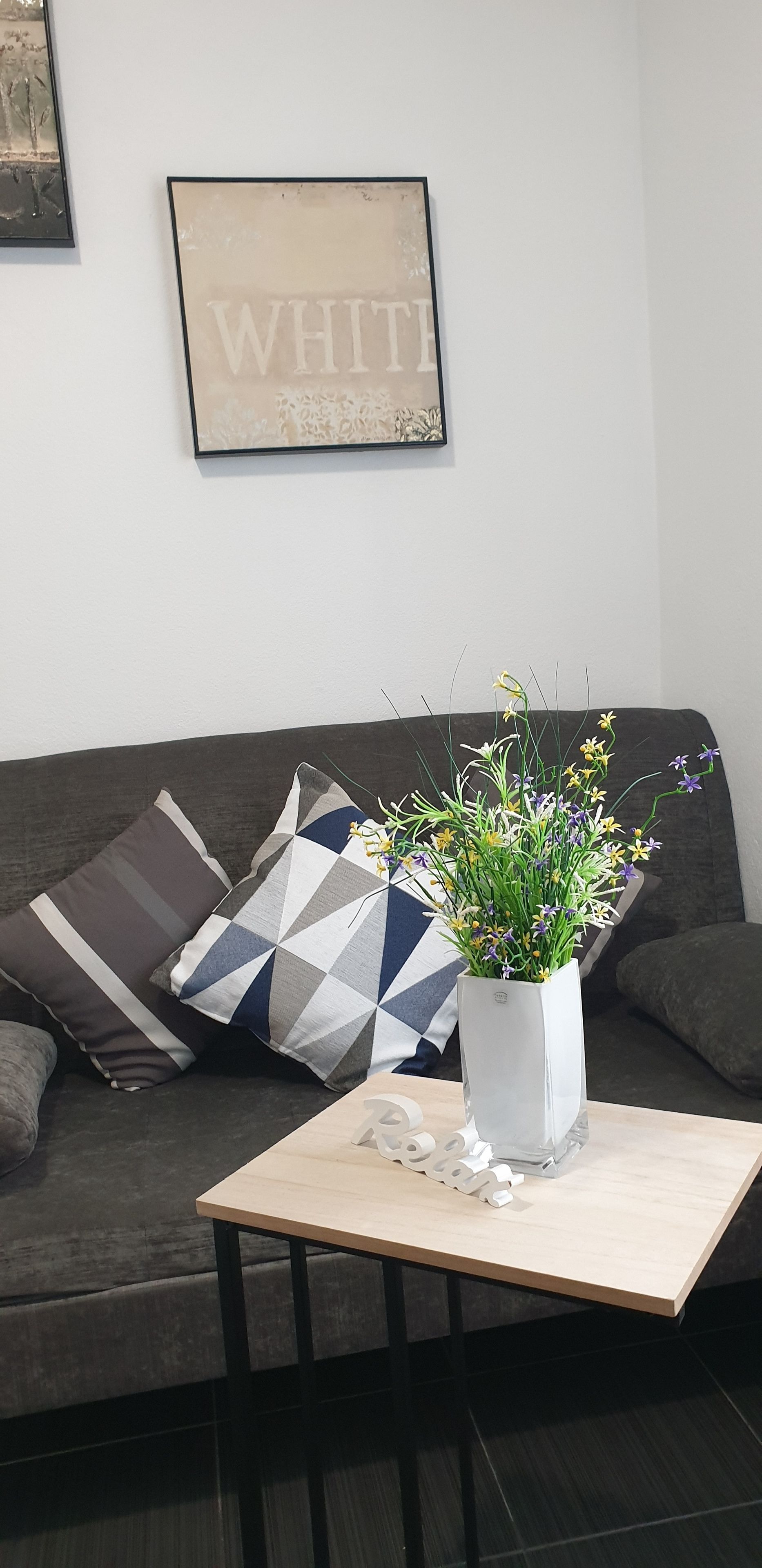 Un salon avec un canapé gris, des coussins à motifs, un vase blanc rempli de fleurs et un tableau avec un mot écrit accroché au mur.