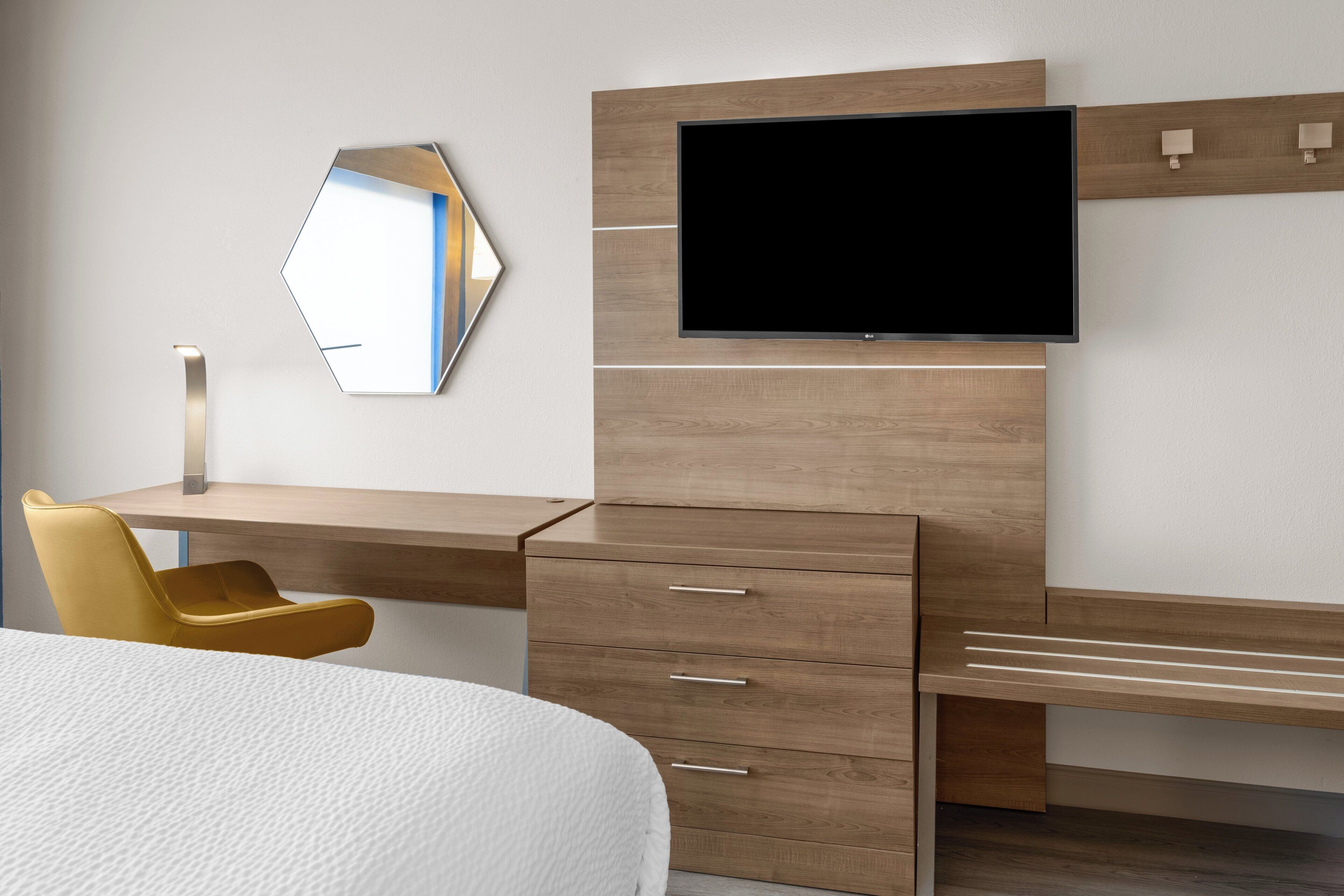 A modern hotel room with a wooden desk, a flat-screen TV, a yellow chair, and a large bed.