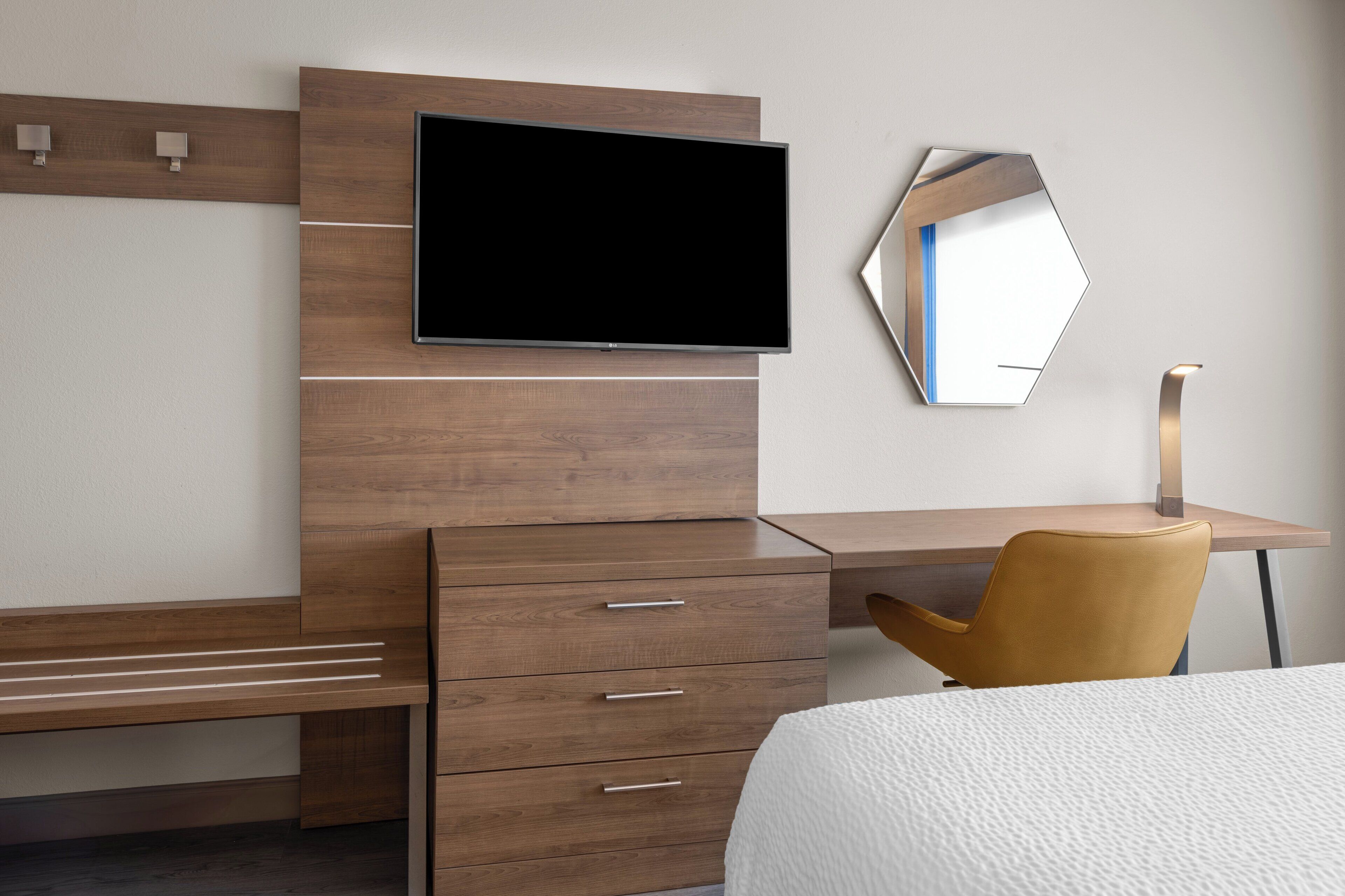 A modern hotel room with a wooden desk, a flat-screen TV, a mirror, and a comfortable chair.