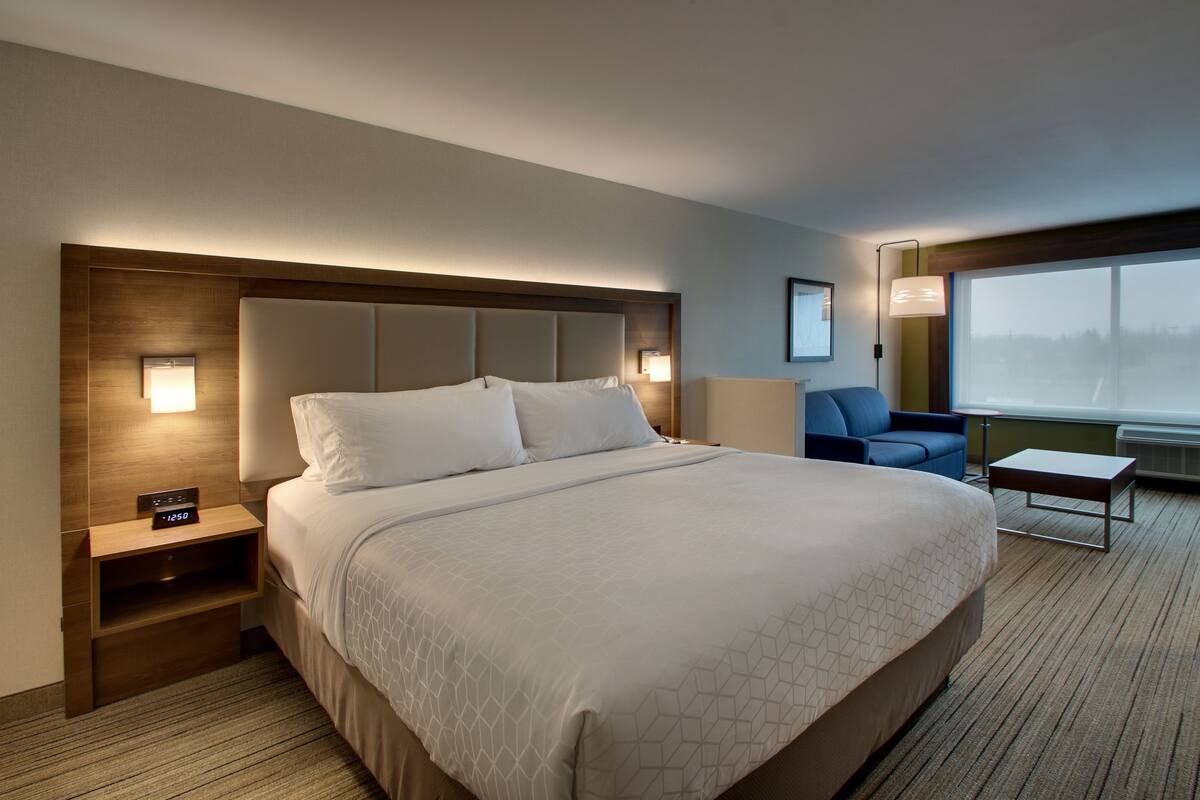 Ein Hotelzimmer mit einem großen Bett, einem Nachttisch mit einer Uhr, einer Sitzecke mit einem Sessel und einem Tisch und einem großen Fenster mit Blick.