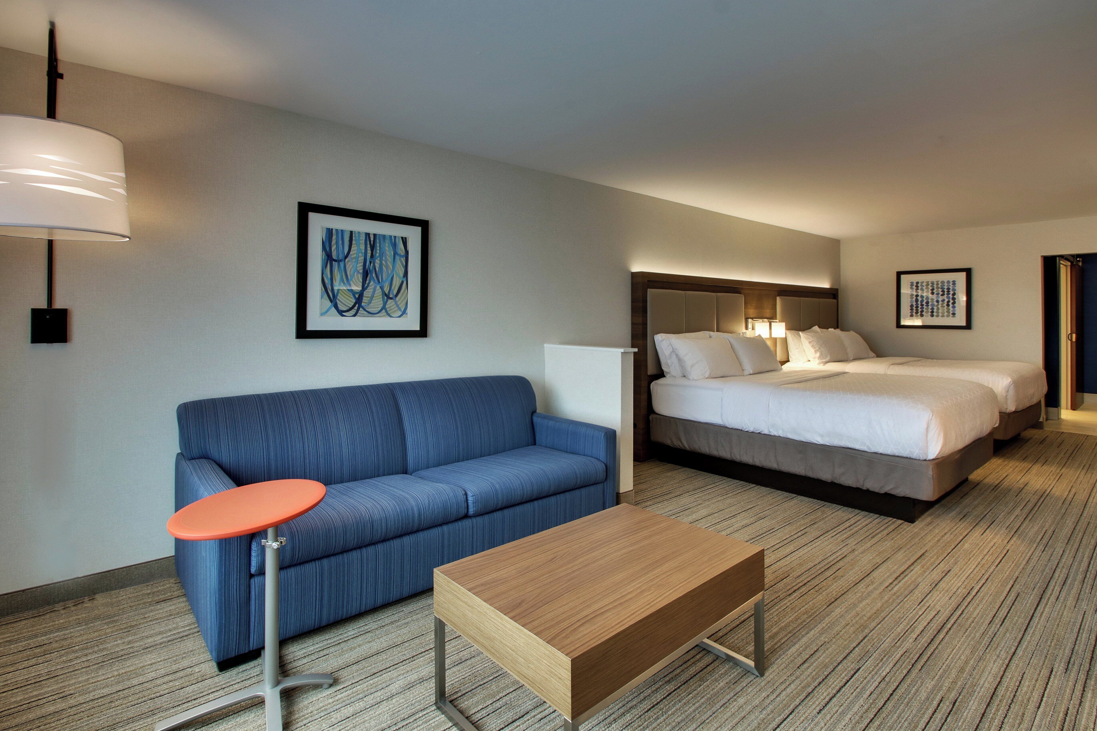 A hotel room with a blue sofa, a wooden coffee table, a bed with white bedding, and two framed artworks on the walls.