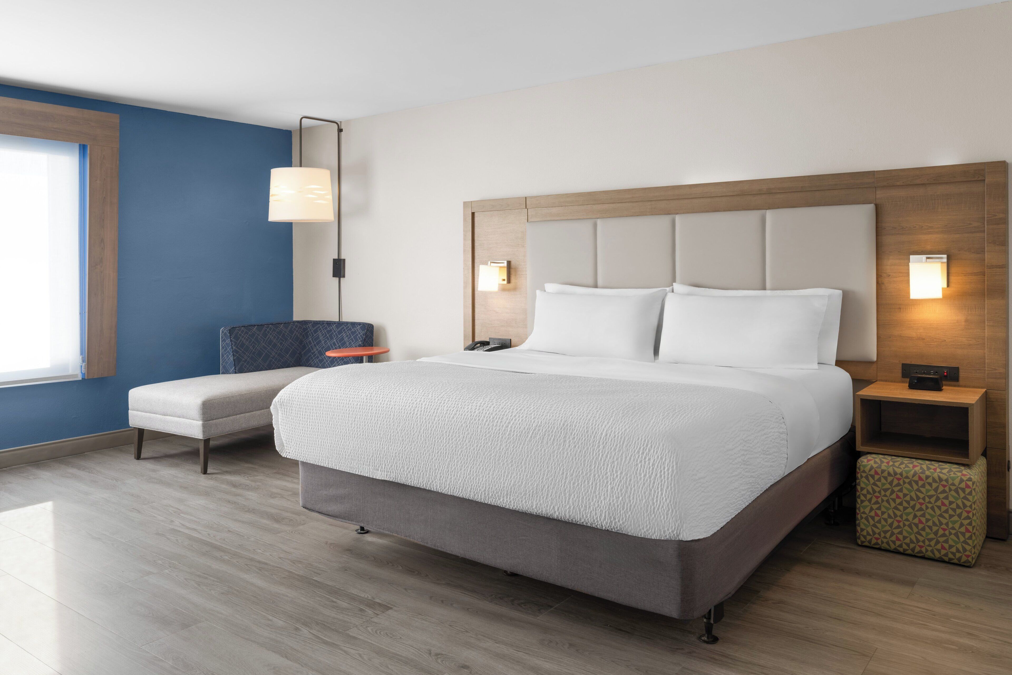 A modern hotel room with a large bed, a bedside table, a chair, and a small ottoman.