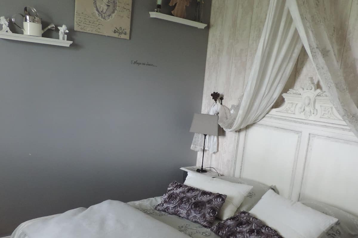 A bedroom with a bed, a nightstand, a lamp, a shelf with decorative items, and a wall with a framed picture.