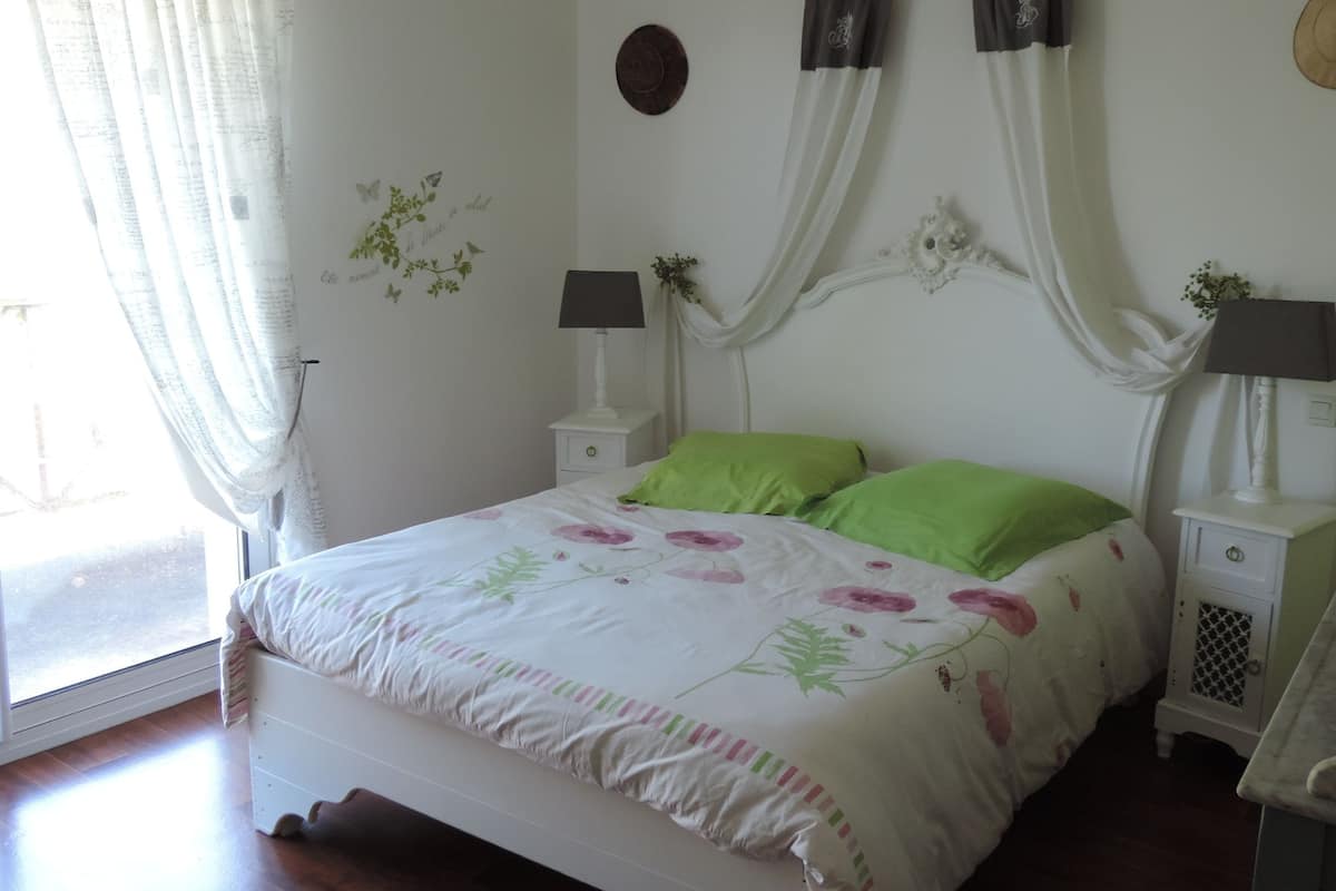 A bedroom with a bed, green pillows, two bedside tables with lamps, a window with curtains, and wall decorations.