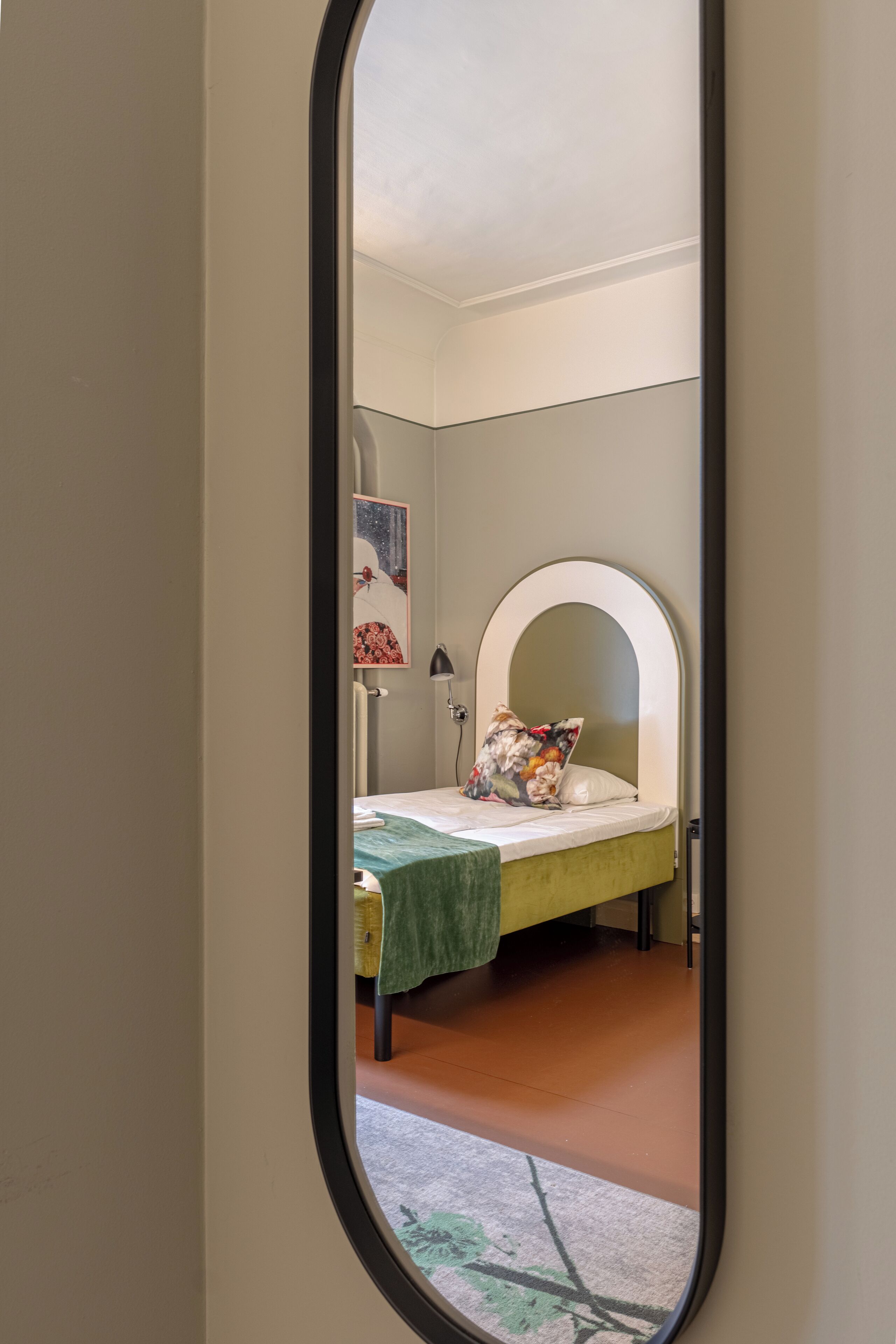 Un dormitorio con una cama, una colcha verde, un almohadón, una lámpara de pared y un cuadro enmarcado en la pared.