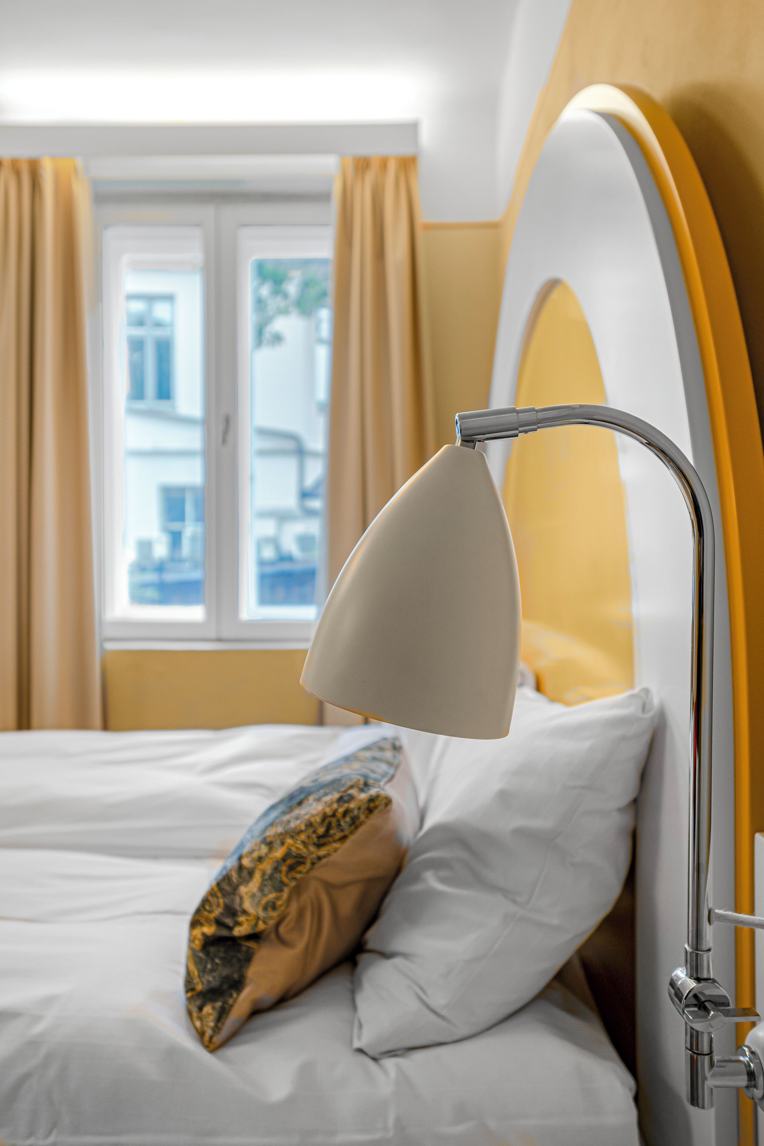 A neatly made bed with a white and patterned pillow, a modern bedside lamp, and a window with curtains in a hotel room.