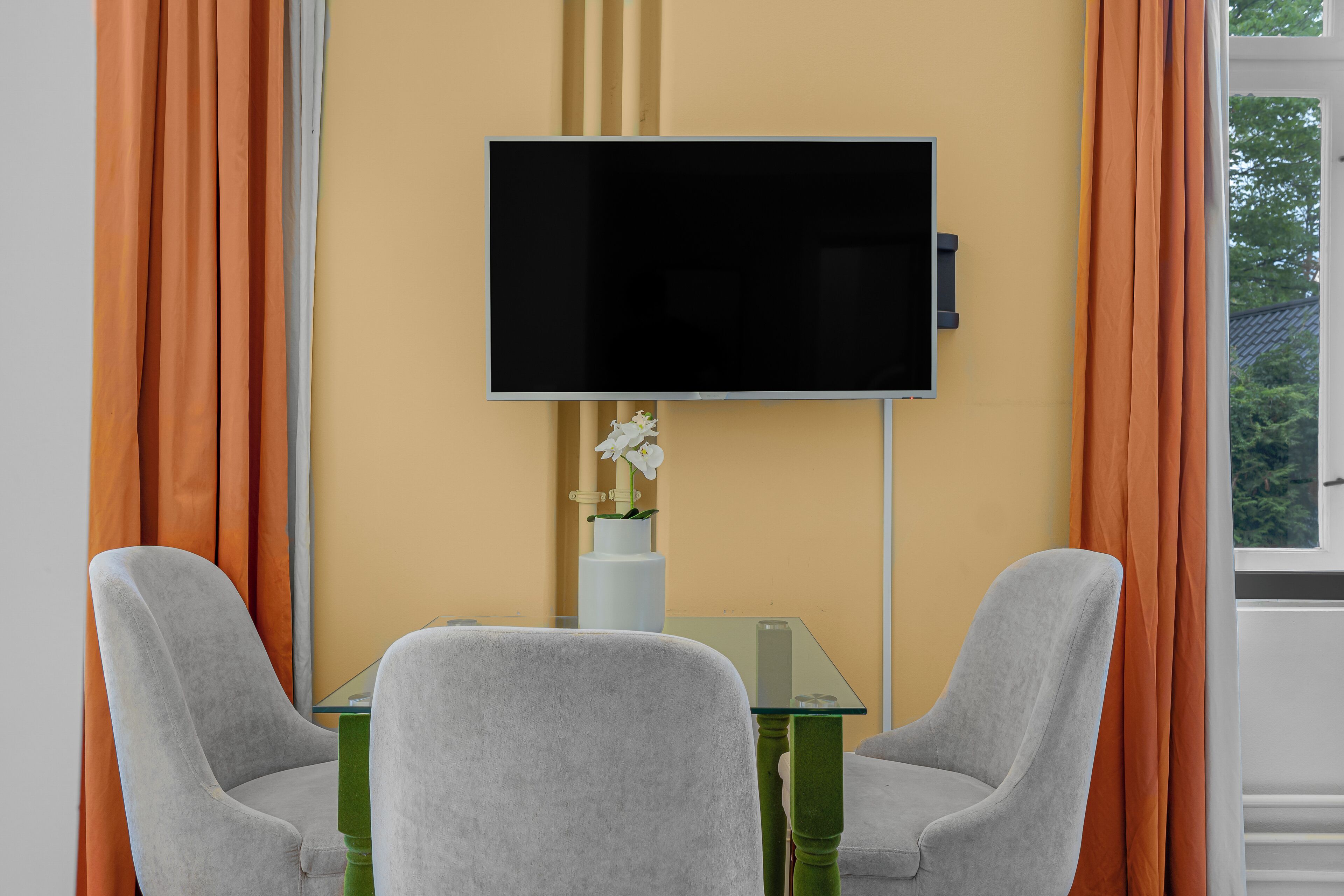 A room with a flat-screen TV mounted on the wall, two grey chairs, a small glass table, and a vase with a white flower.