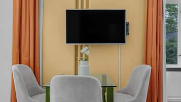 A room with a flat-screen TV mounted on the wall, two grey chairs, a small glass table, and a vase with a white flower.