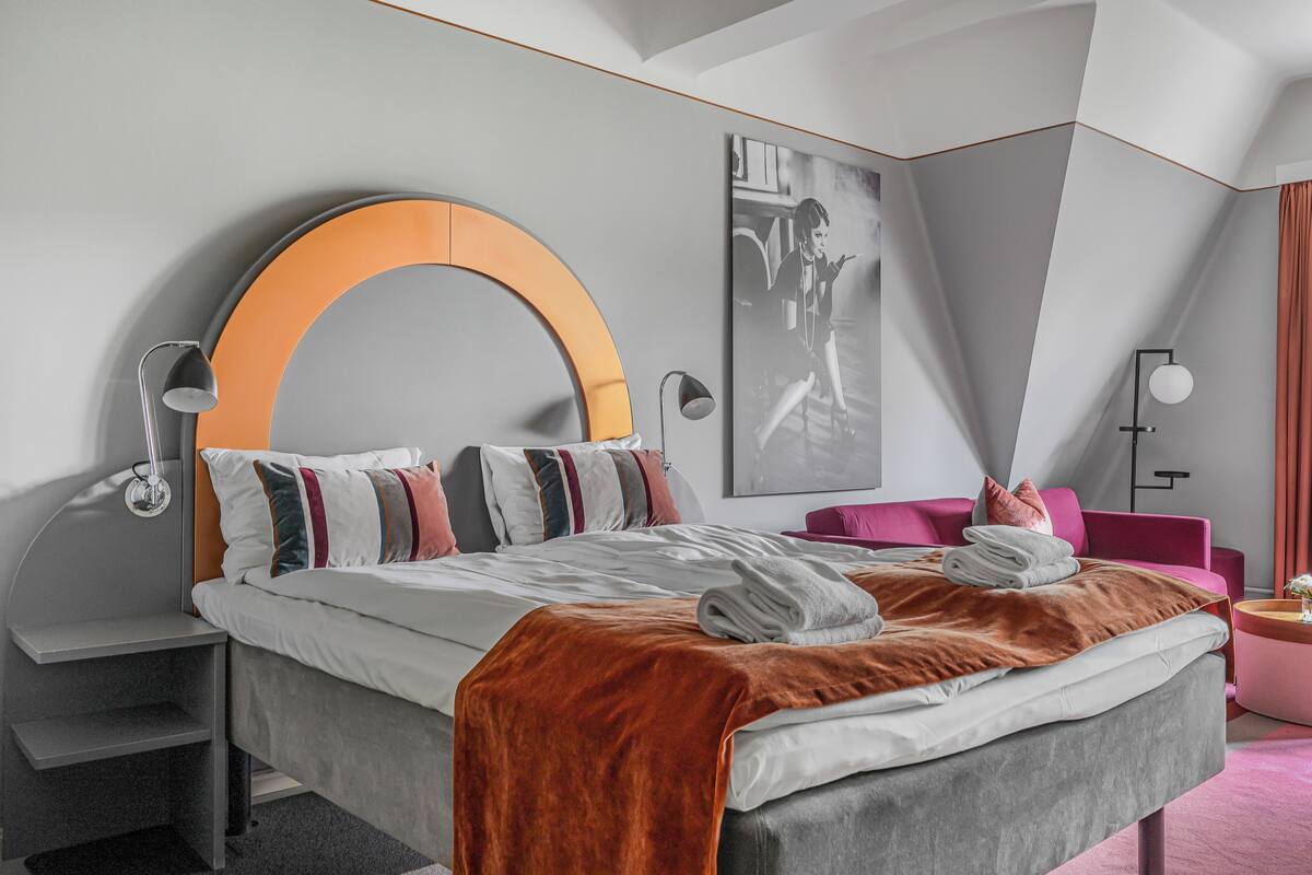 A modern hotel room with a large bed, a curved headboard, a pink armchair, and a wall-mounted artwork.