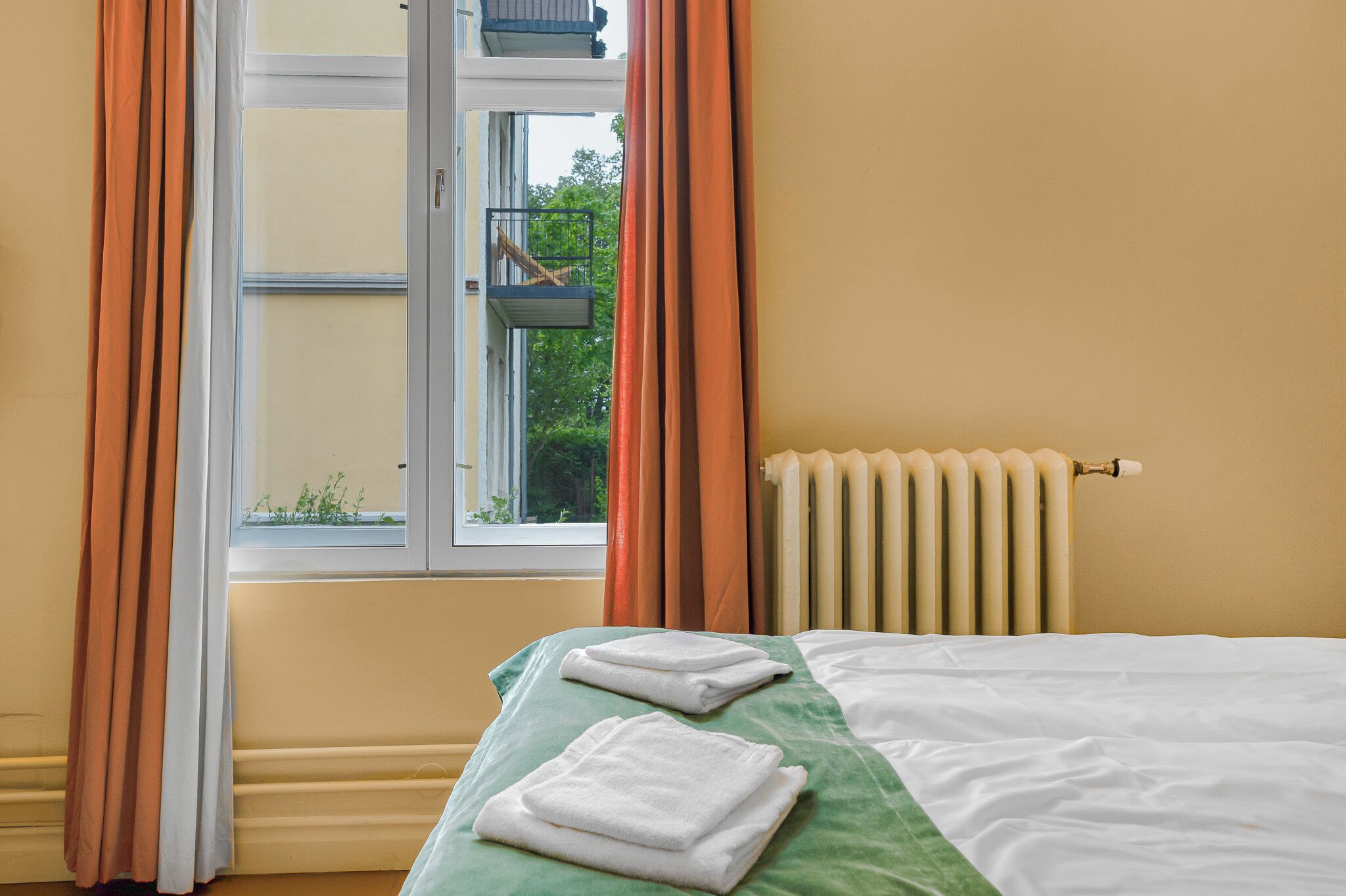 A hotel room with a bed, a window with curtains, a radiator, and a view of greenery outside.