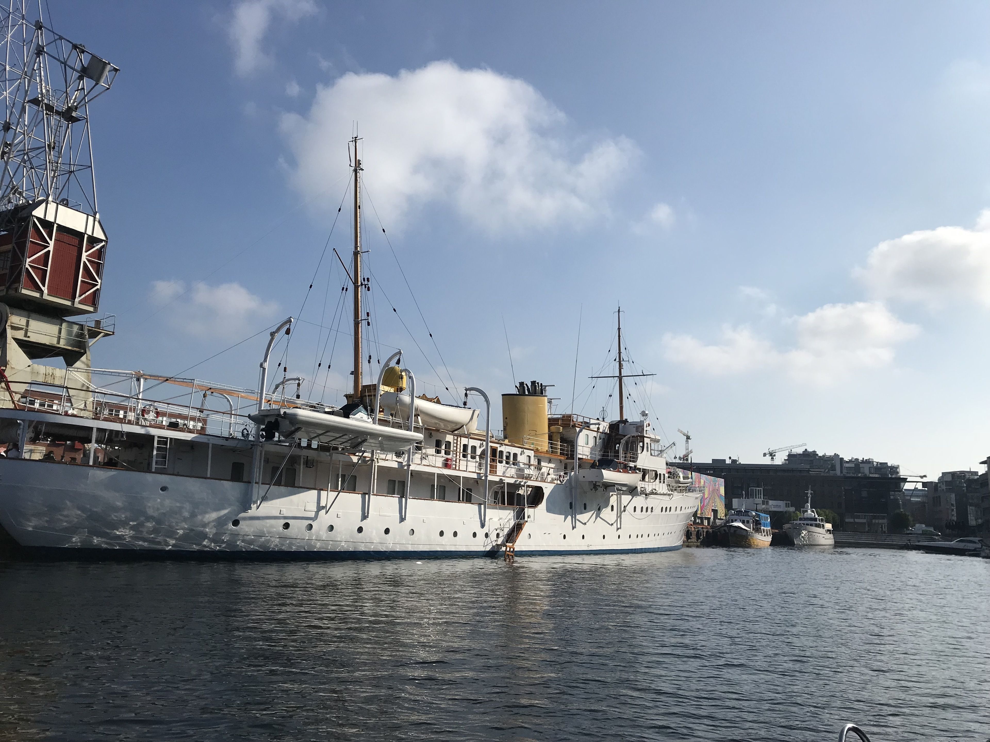 A large white yacht docked at a marina with a crane and other boats in the background.